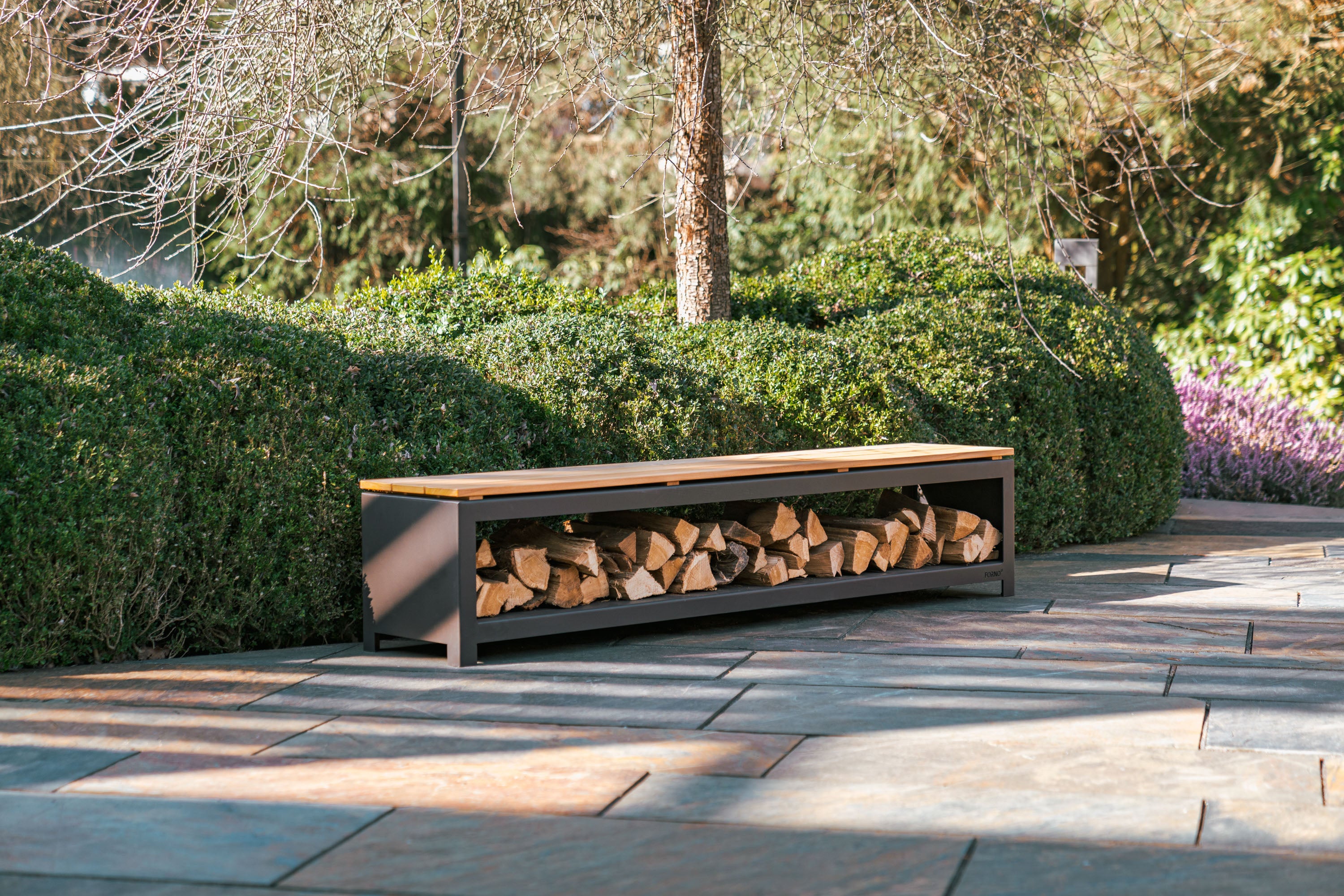 black wood storage bench with wooden top filled with fire wood on paving next to hedge