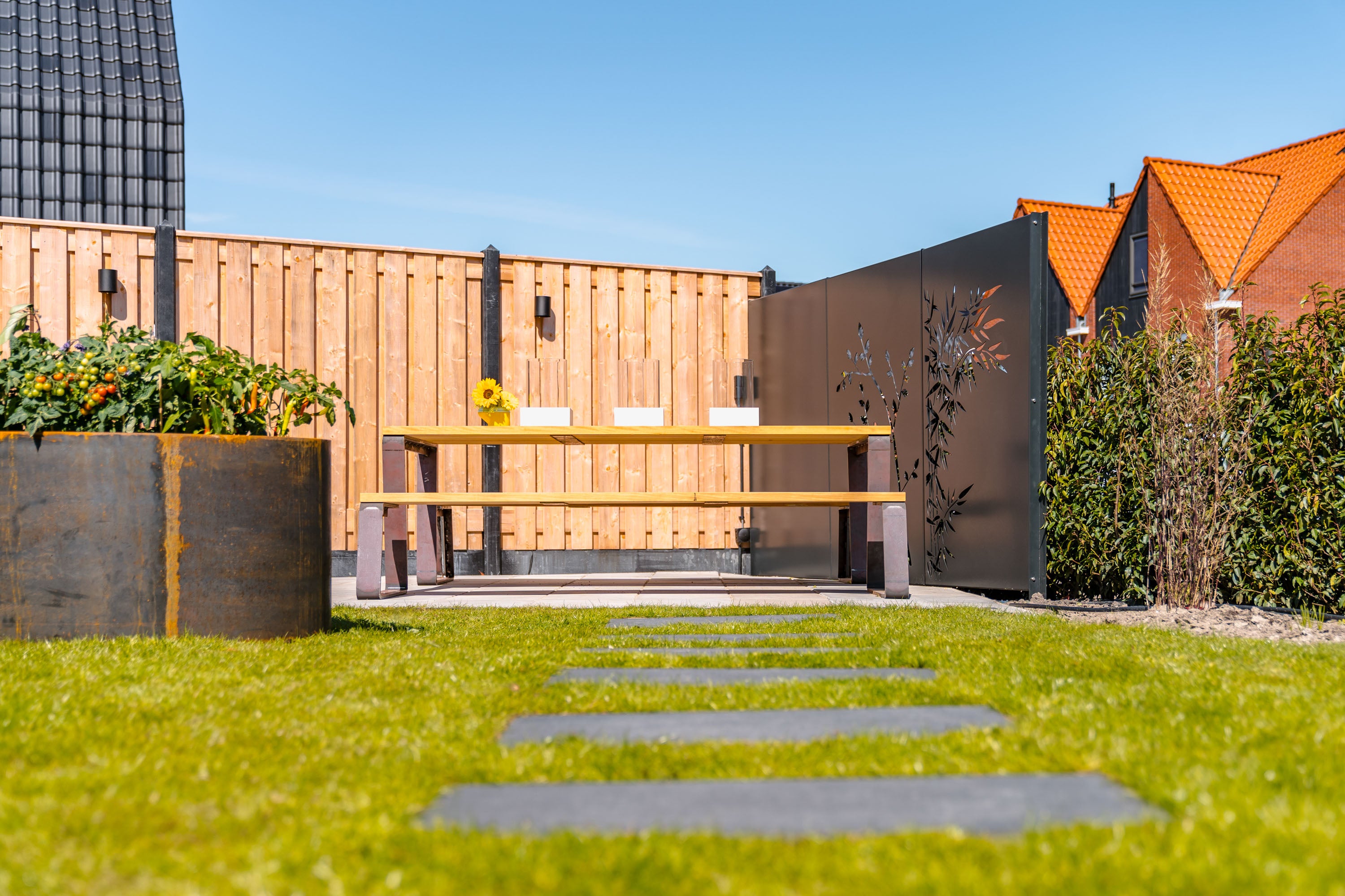Modern outdoor patio scene with a wooden fence, dark metal wall panels, a wooden dining table and benches, a large planter with greenery, a grassy lawn with stone pavers, and neighboring houses under a blue sky.