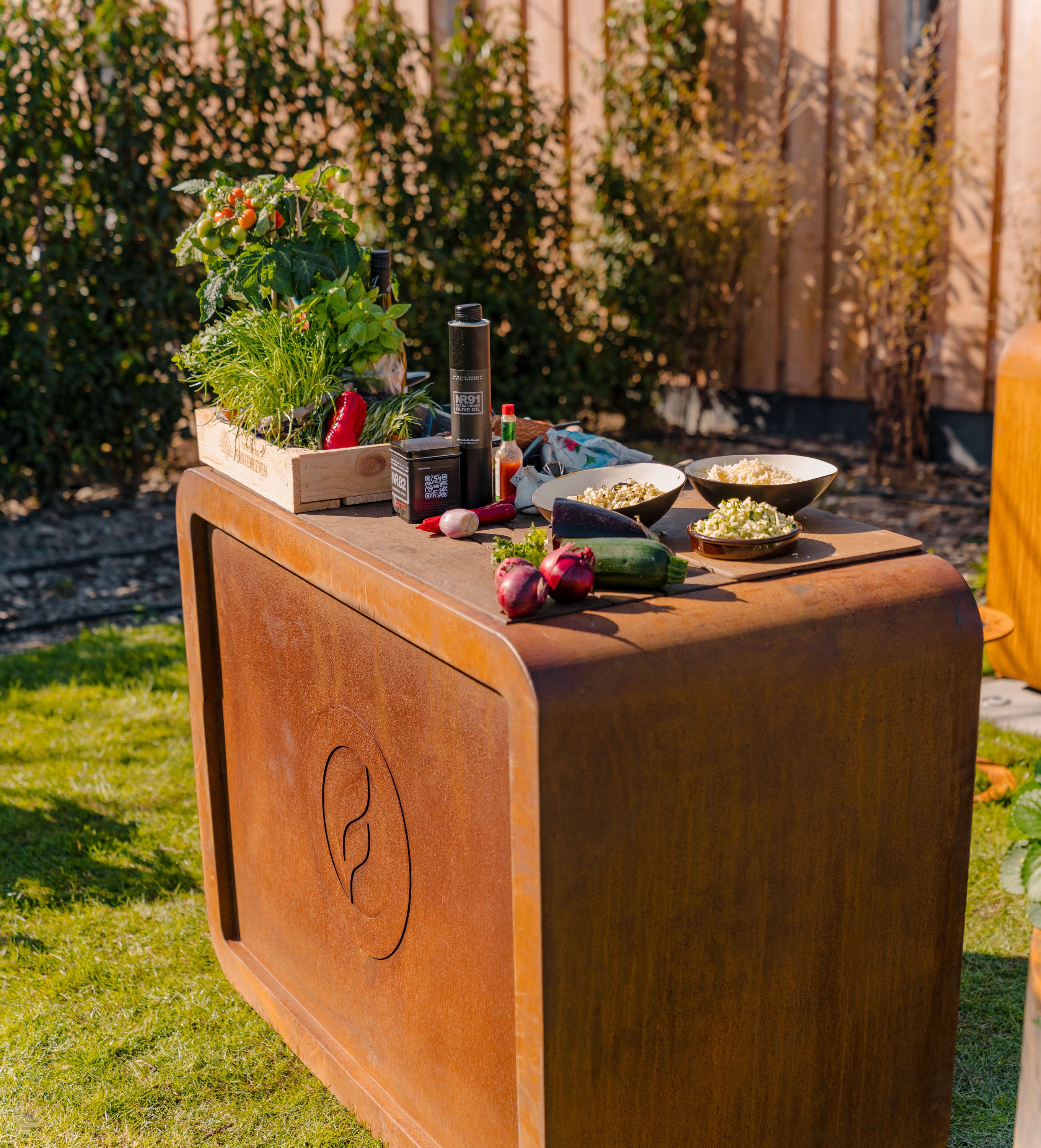 Outdoor setting with a wooden table displaying fresh produce and a canister, surrounded by greenery.