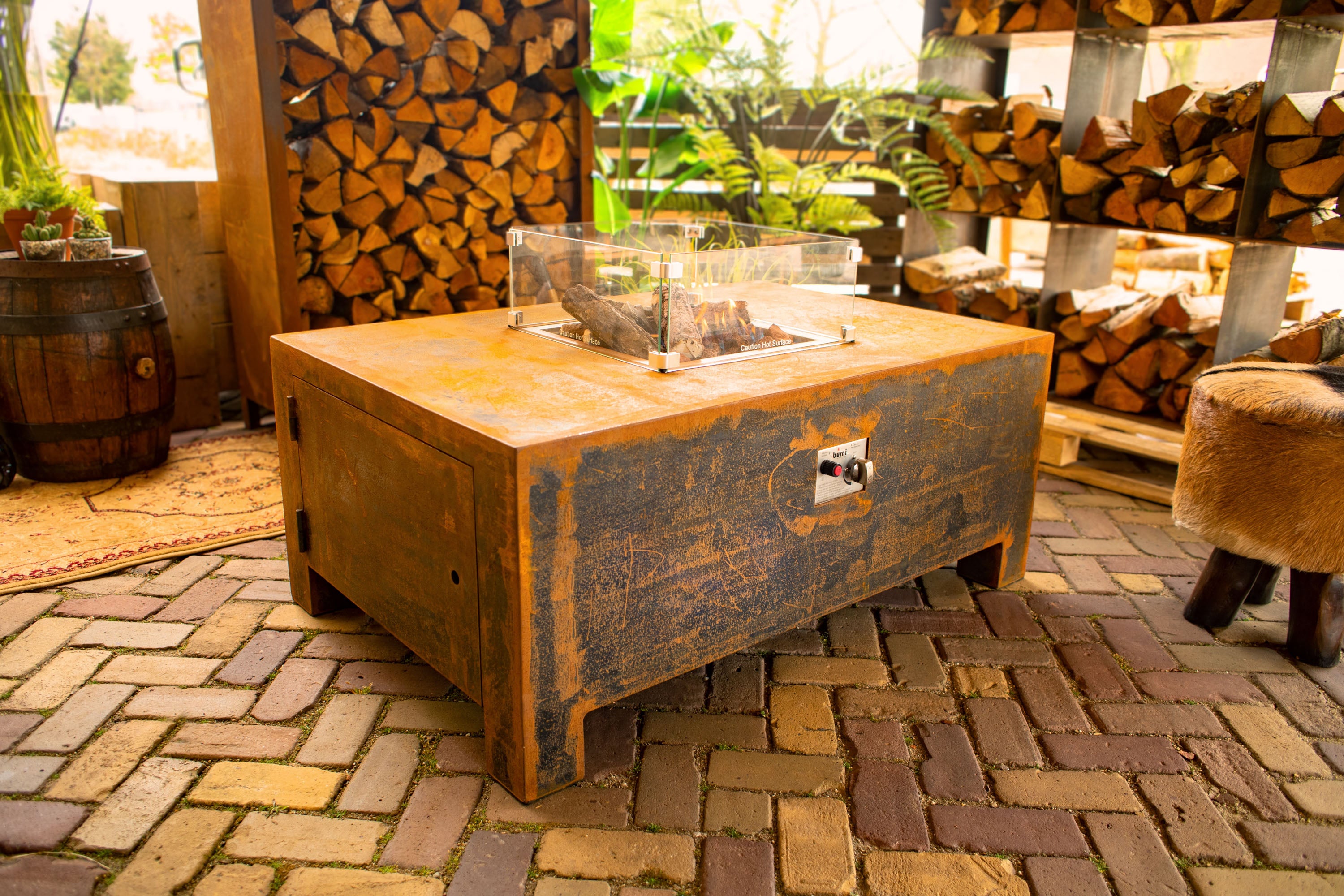 rusted fire table with clear central screen around imitation logs. situated  on brick paving next to a wood store