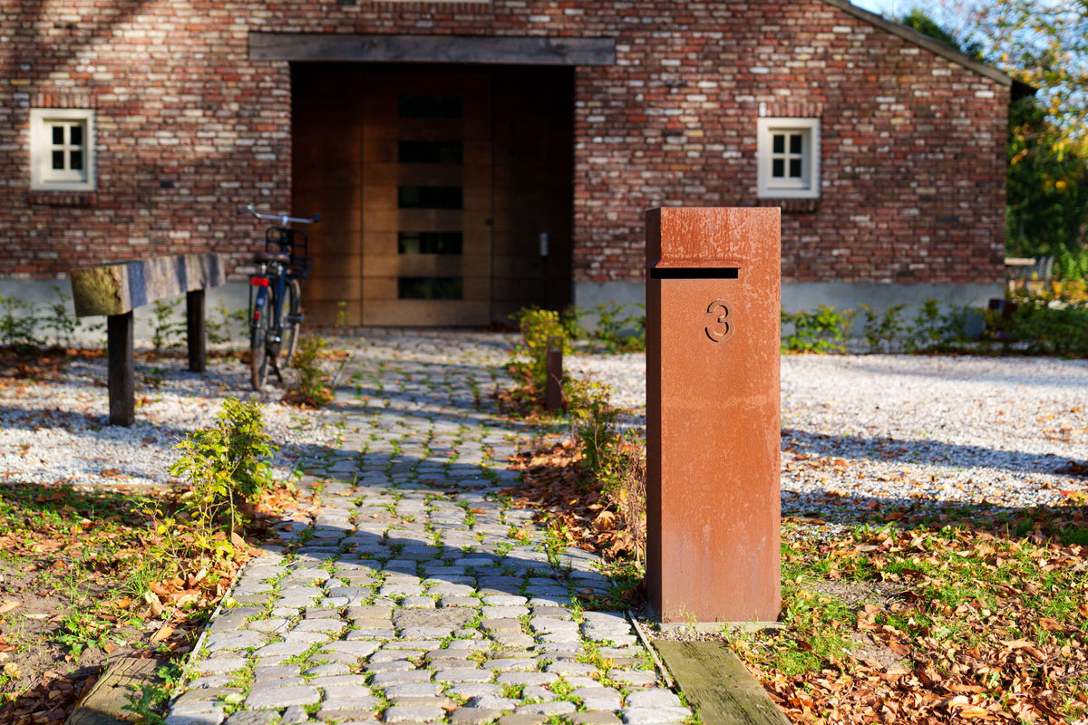 A rust-colored metal mailbox with a slotted top stands on a cobblestone path in front of a brick building with a wooden garage door and a bicycle nearby.