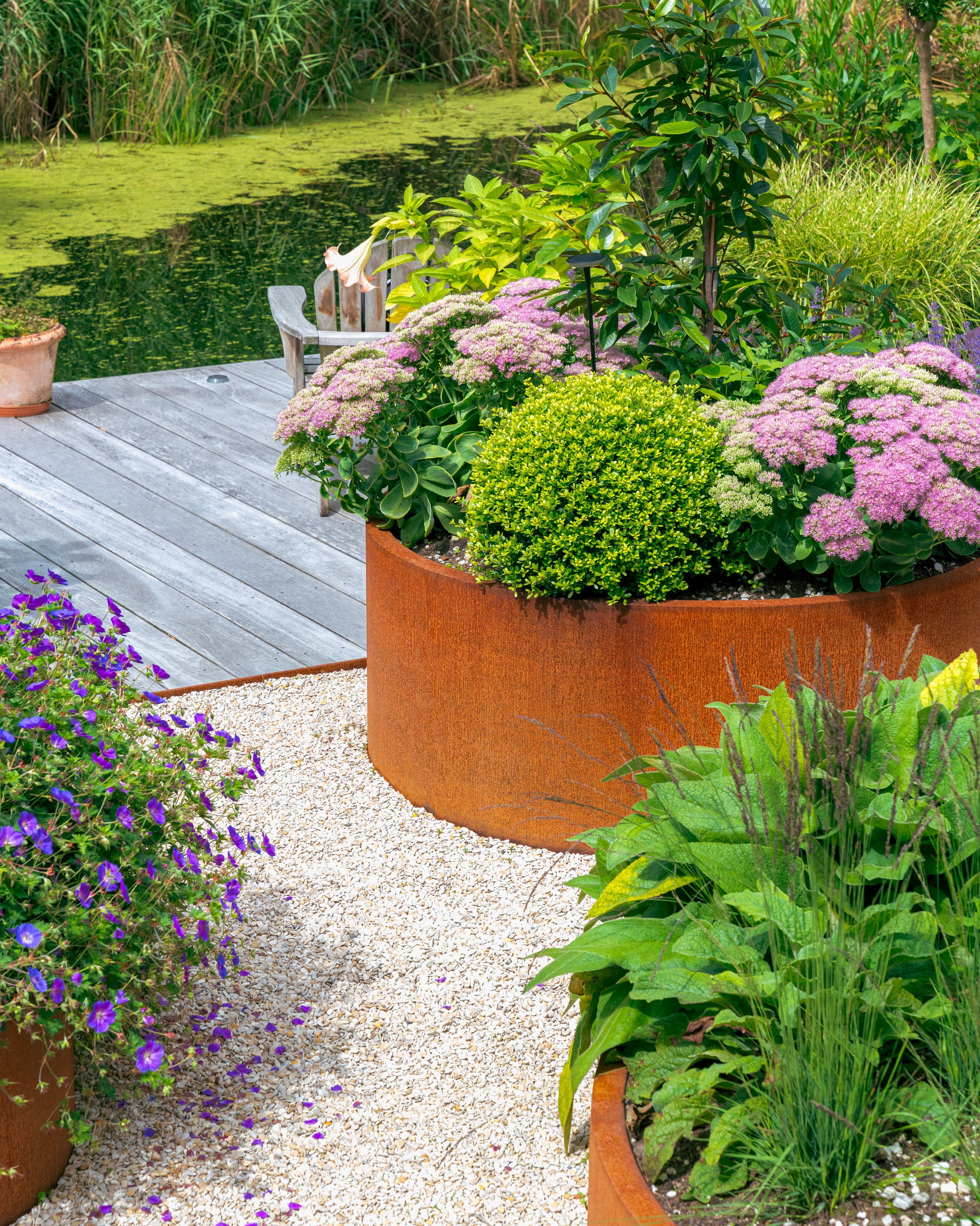 Garden scene with a round metal planter filled with plants and flowers by a pond.