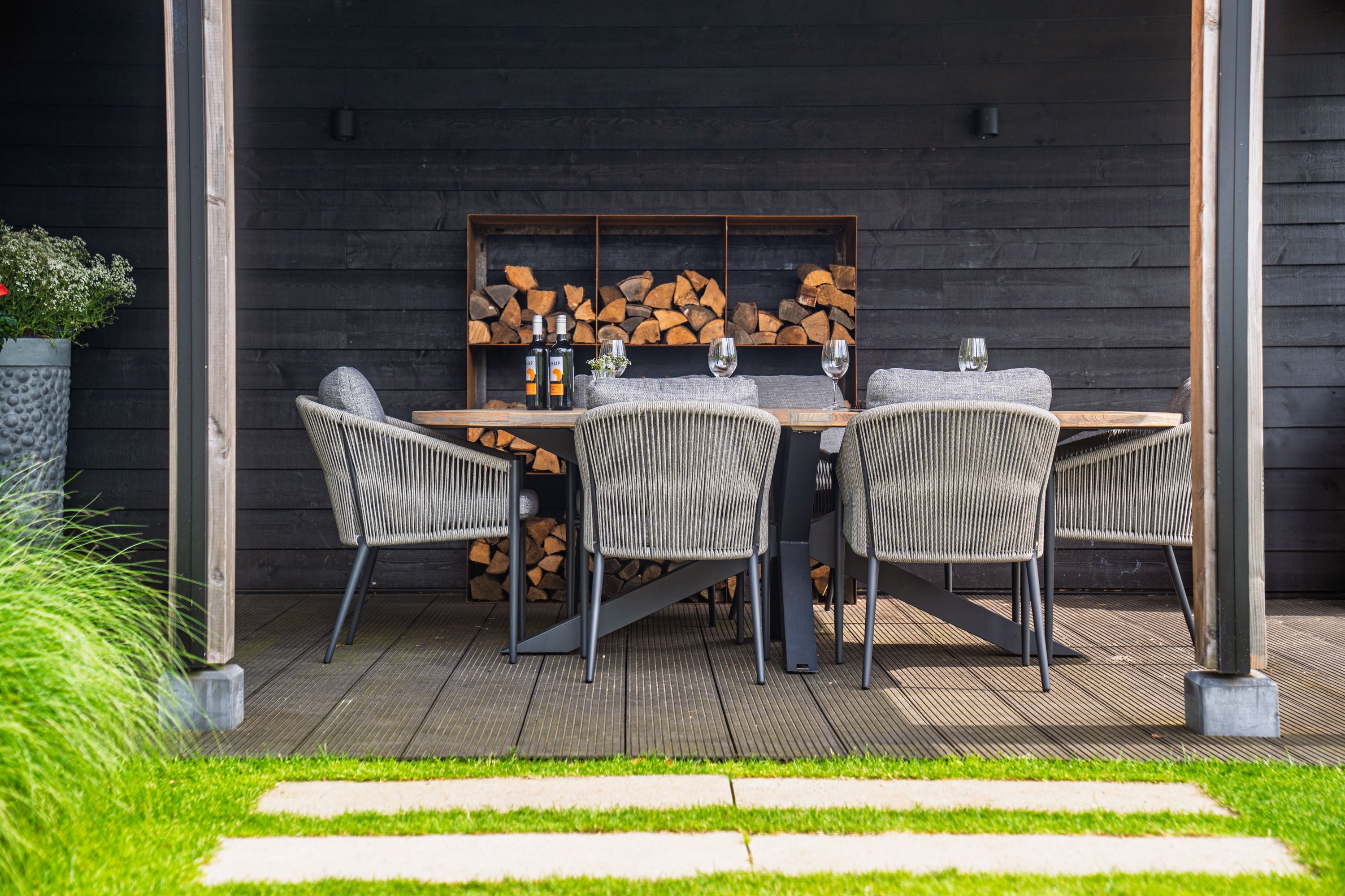 Outdoor dining area with a wooden table and chairs in front of a dark wooden wall with firewood storage.