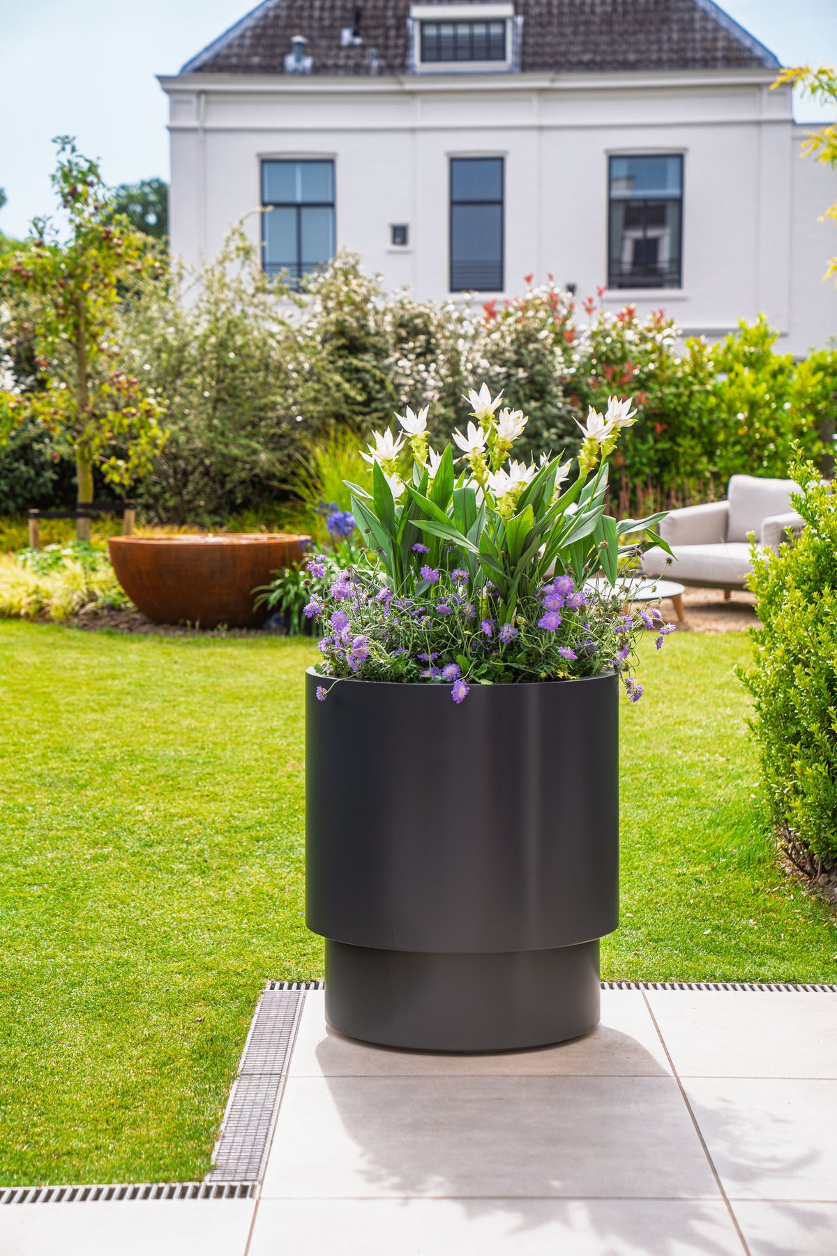 A modern dark gray cylindrical planter filled with white and purple flowers, set on a tiled patio in a lush green backyard with a house in the background.