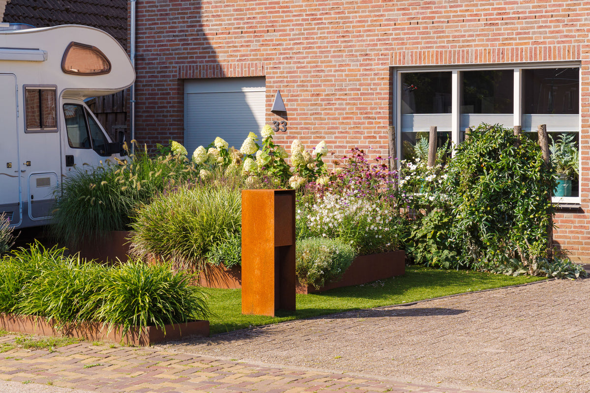 A modern rust-orange rectangular steel planter sculpture stands on a green lawn in a landscaped garden in front of a brick house. The scene includes potted plants, a brick pathway, and a white motorhome on the left.