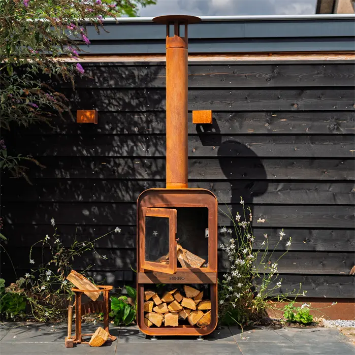 A rusted orange outdoor wood-burning stove with a tall chimney pipe, shown standing on a patio in front of a dark wooden wall with plants around it.