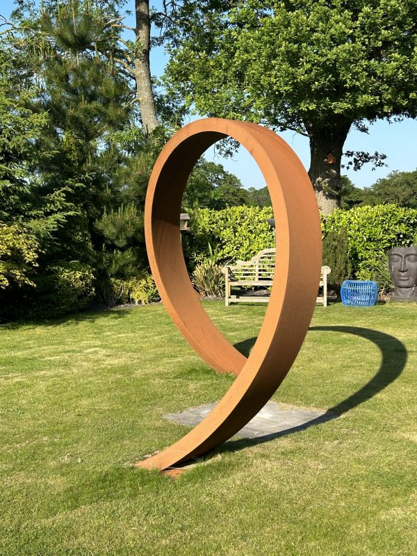 Large corten metal sculpture of two interlocking rings on a grassy area with trees and a bench in the background.