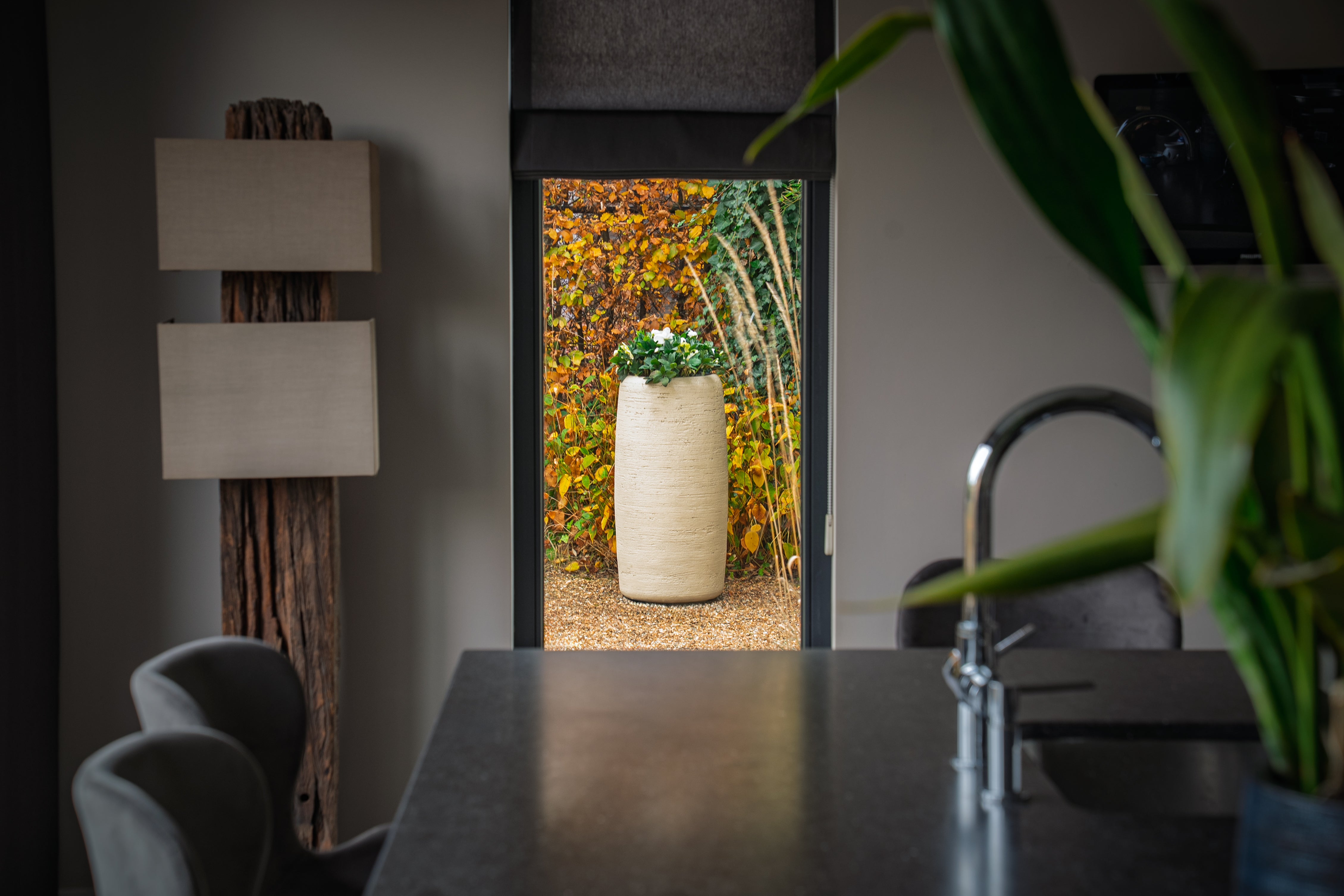 A modern interior scene with a dark countertop, a curved faucet, gray chairs, a tall floor lamp, and a large green plant in the foreground, framing a view of a planter with autumn foliage outside.