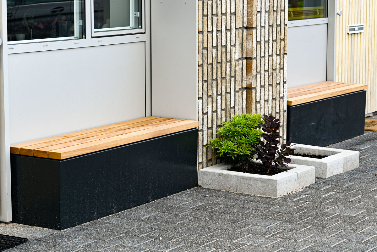 Modern outdoor wooden bench seating with black metal bases, installed beside a building entrance on a paved area with a planter in between.