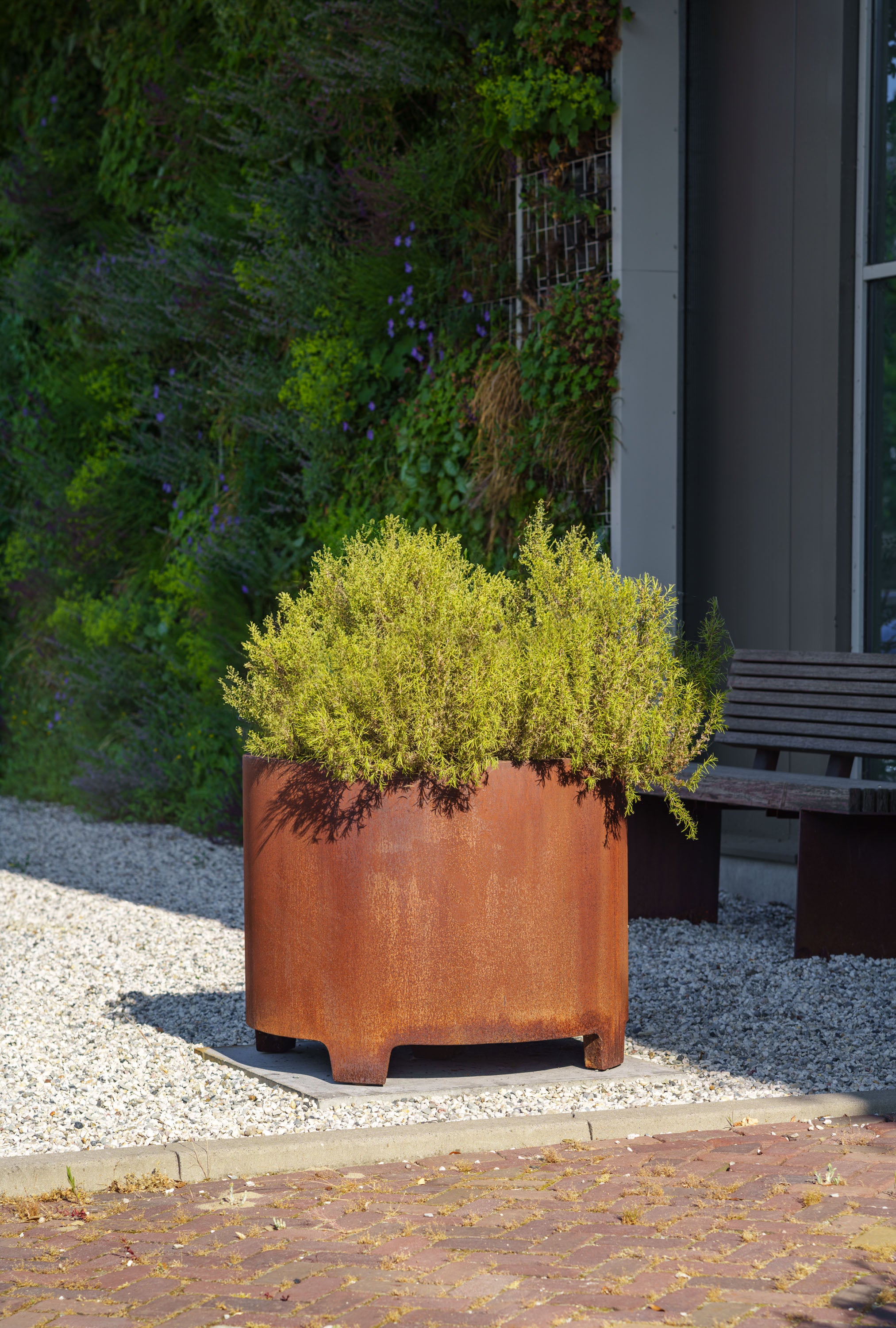 Rust-colored planter with green plants on a pebbled ground next to a building.