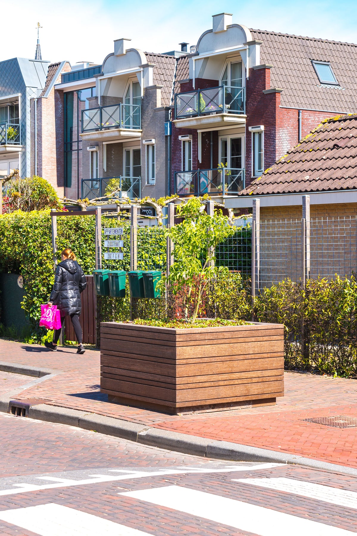 A large rectangular wooden planter box filled with green plants sits on a red brick sidewalk in front of modern multi-story residential buildings. A person carrying a bright pink bag walks along the sidewalk in the background.