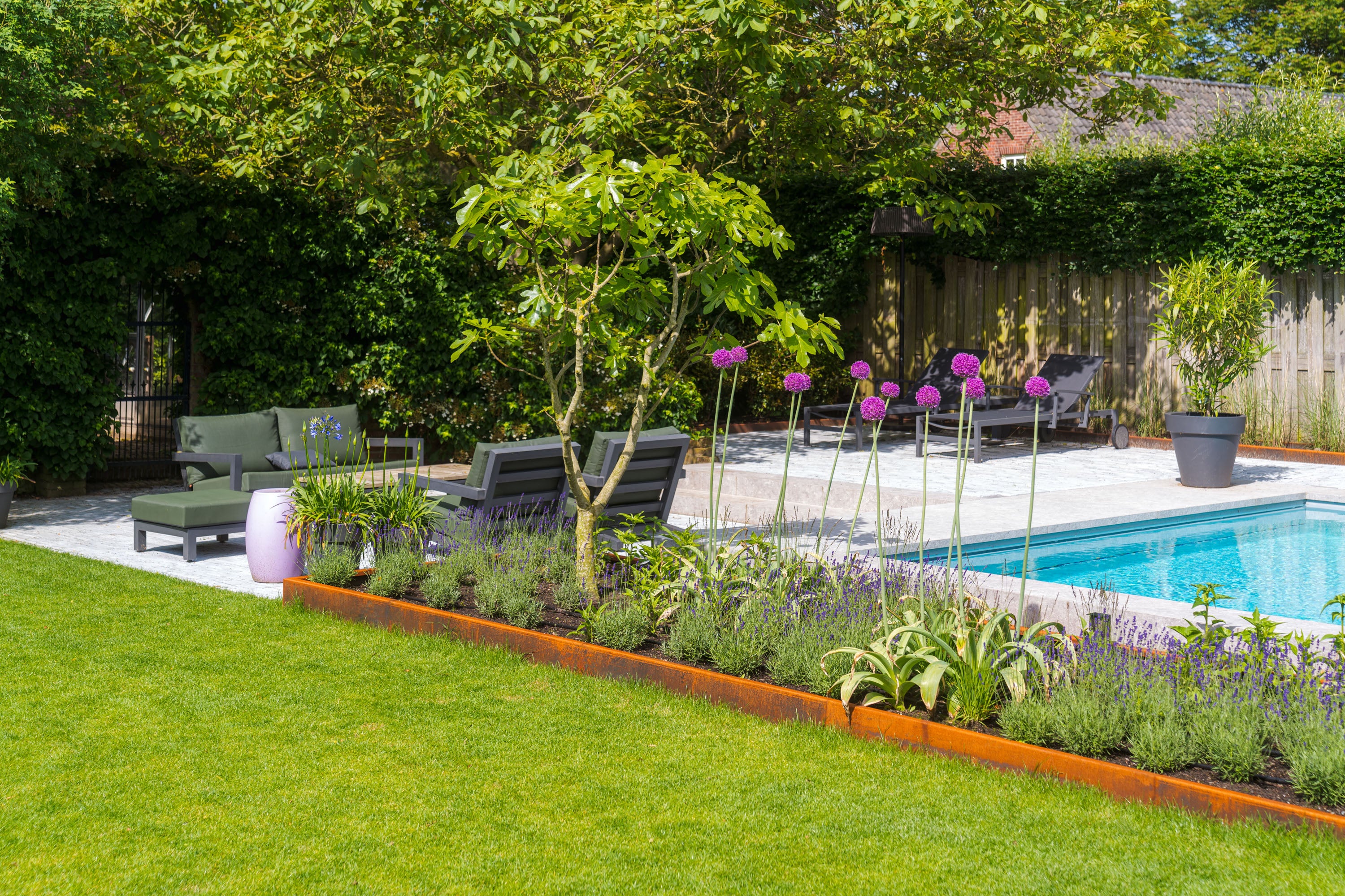 A landscaped backyard garden with a swimming pool, outdoor lounge chairs, a green sofa, potted plants, and a wooden fence, with a prominent raised corten garden bed in the foreground.
