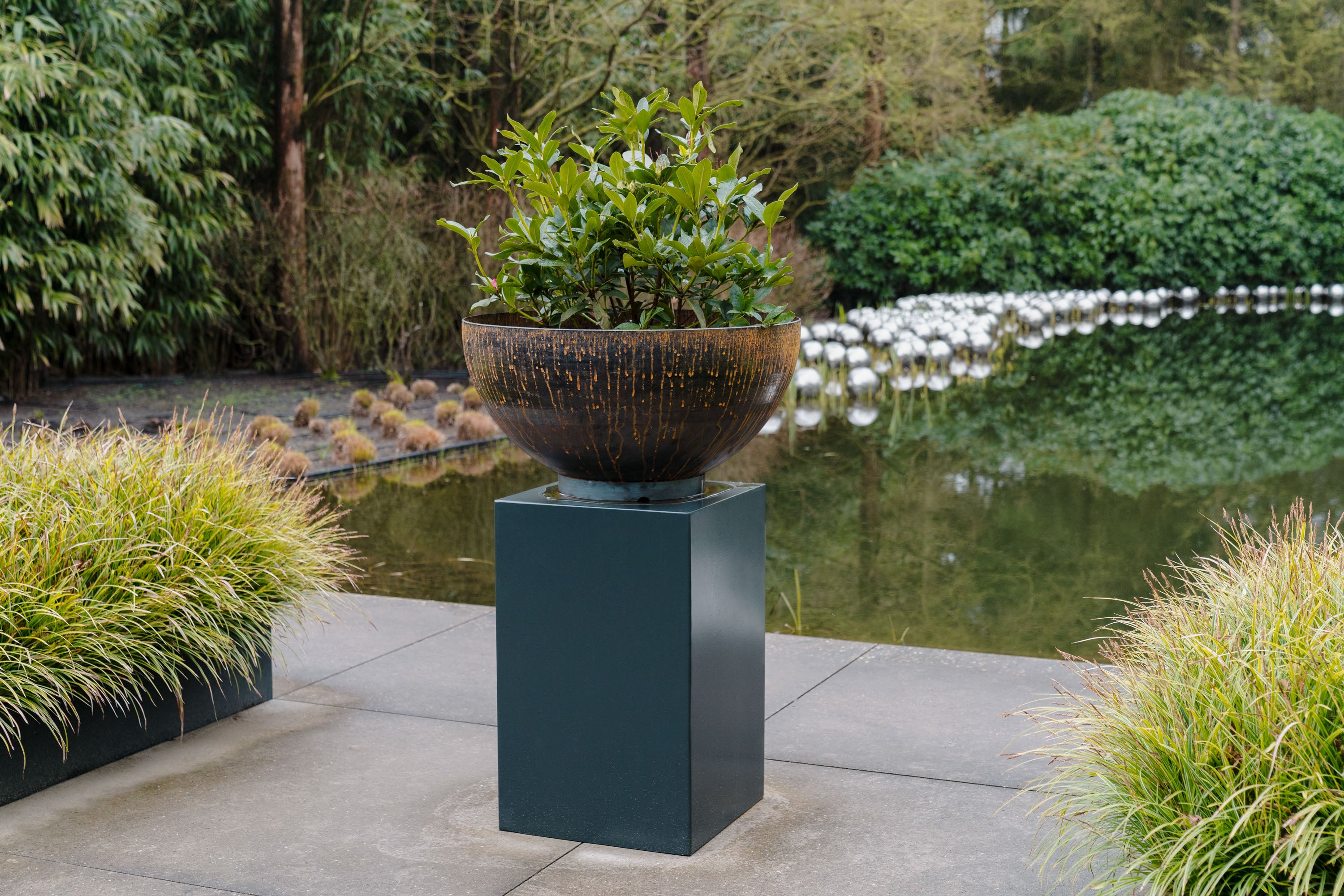 Decorative planter on a pedestal in a garden setting with a pond and greenery.