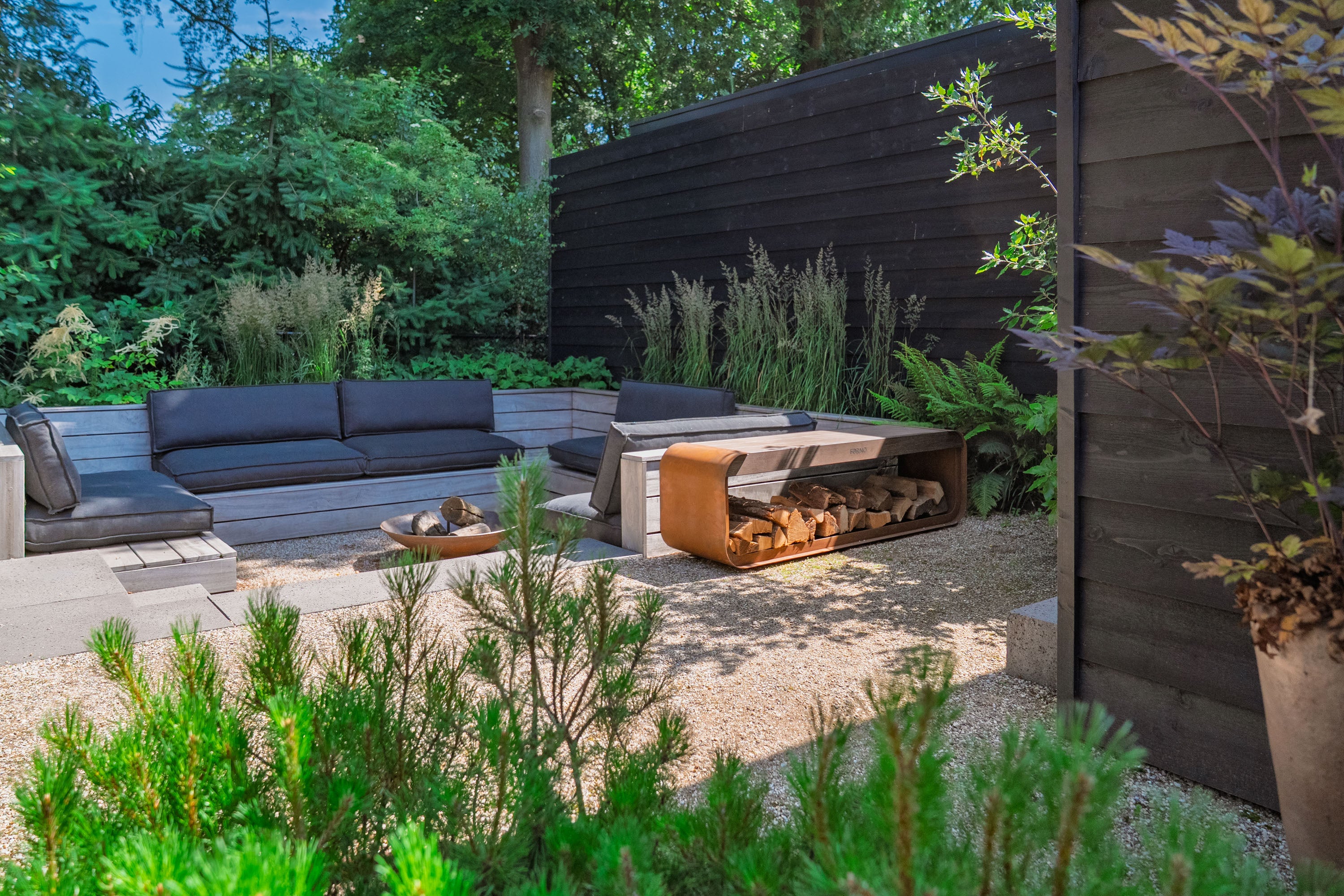 Modern outdoor patio furniture set in a landscaped backyard, featuring a dark gray sectional sofa, a low concrete fire pit with wood logs, and a matching concrete coffee table, surrounded by green plants and a dark wooden fence.