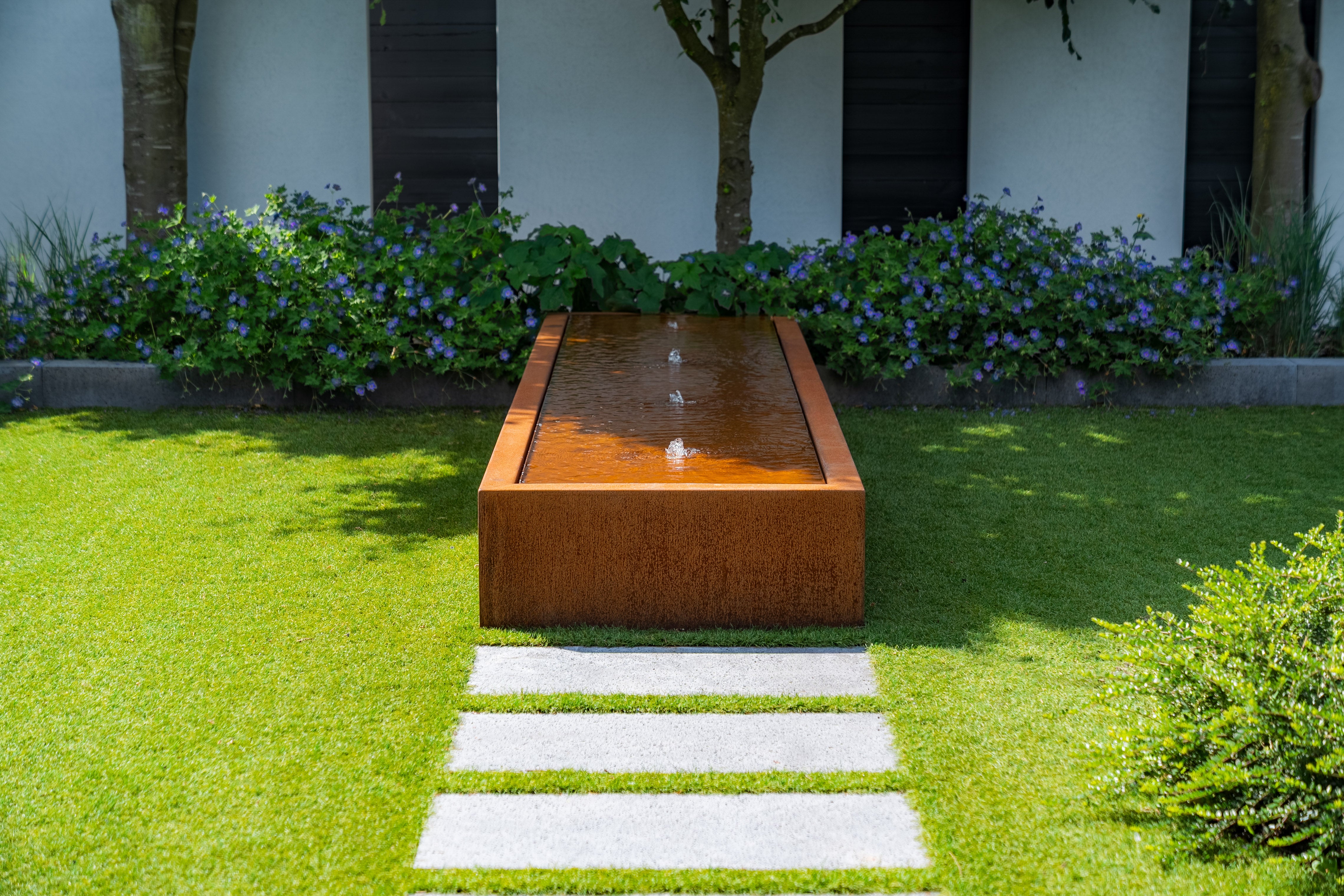 rusted water table with fountains in the middle, filled with water on grass next to bed planted with flowers