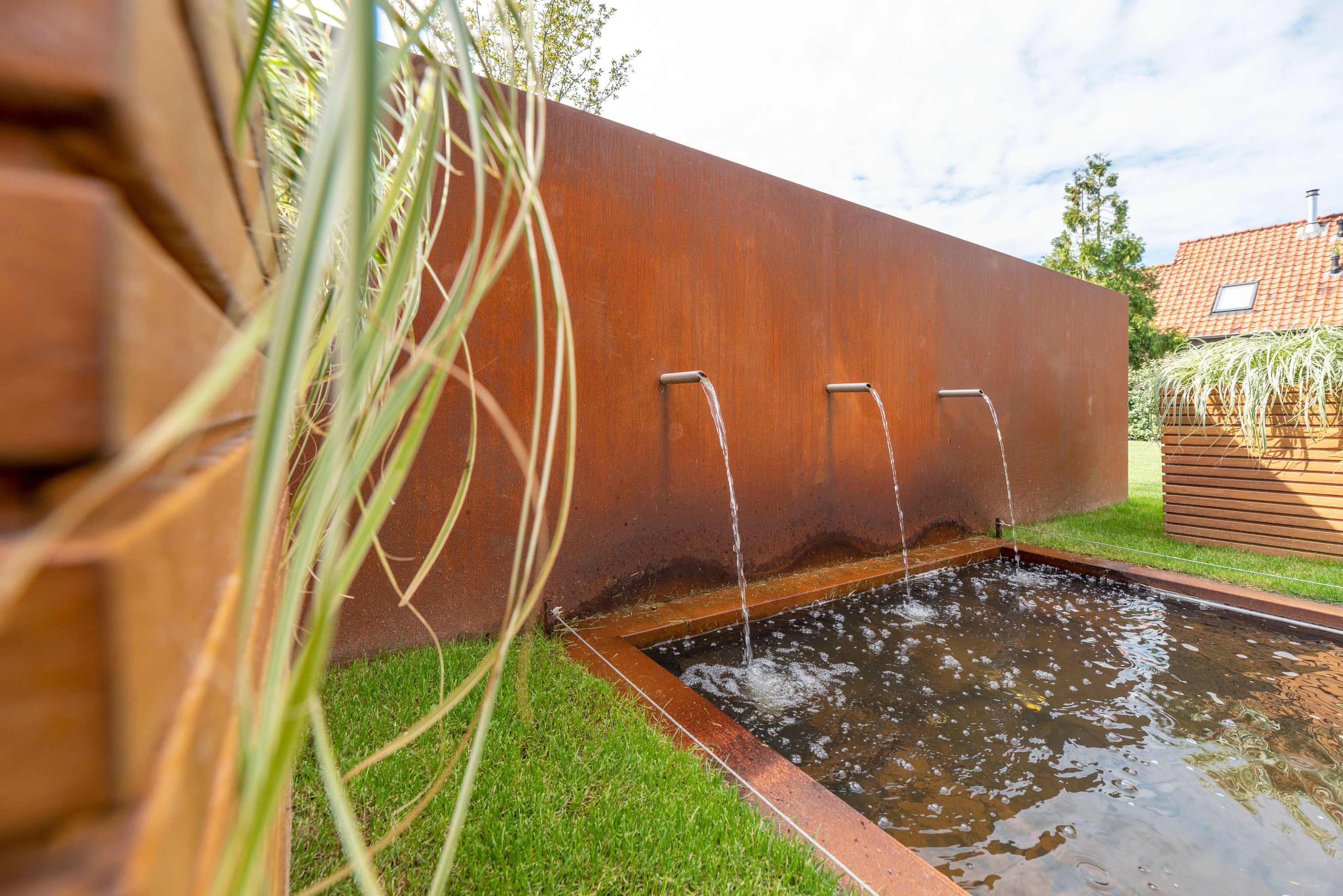 A modern outdoor water feature with three metal faucets spraying into a rectangular pool, set against a large rusted corrugated metal wall with grass and a wooden fence in the foreground.