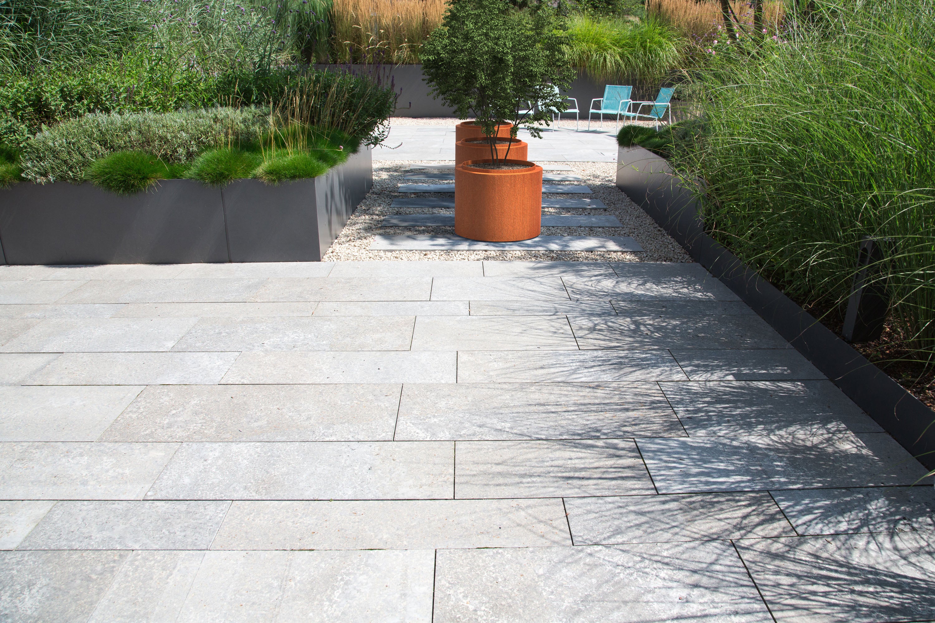 A modern outdoor patio paved with large light gray rectangular stone tiles, bordered by dark gray planter boxes with green ornamental grasses and a central terracotta planter holding a small tree.