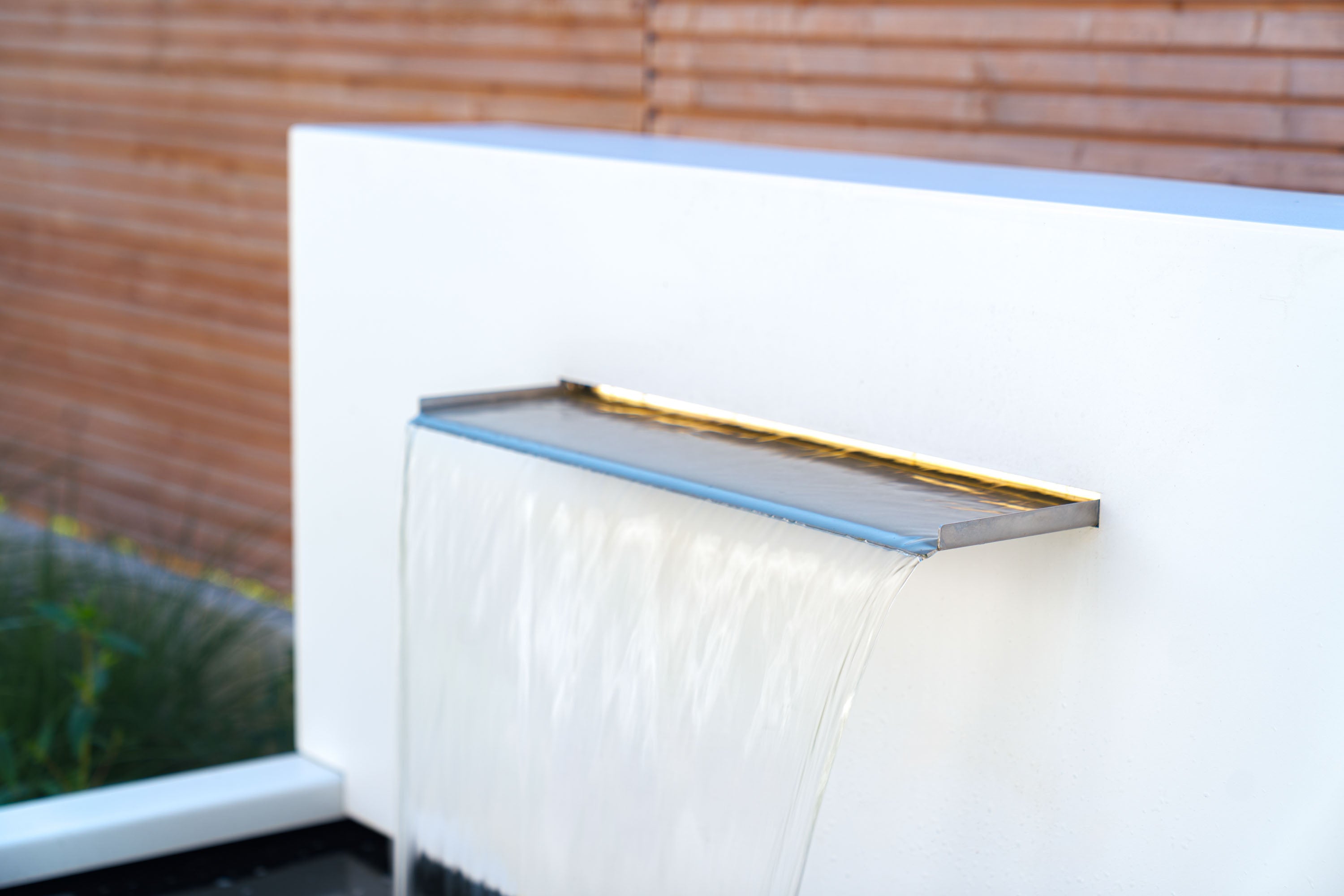 A modern rectangular waterfall wall fountain with a stainless steel tray and warm gold LED lighting, mounted on a white concrete wall with a brick background.