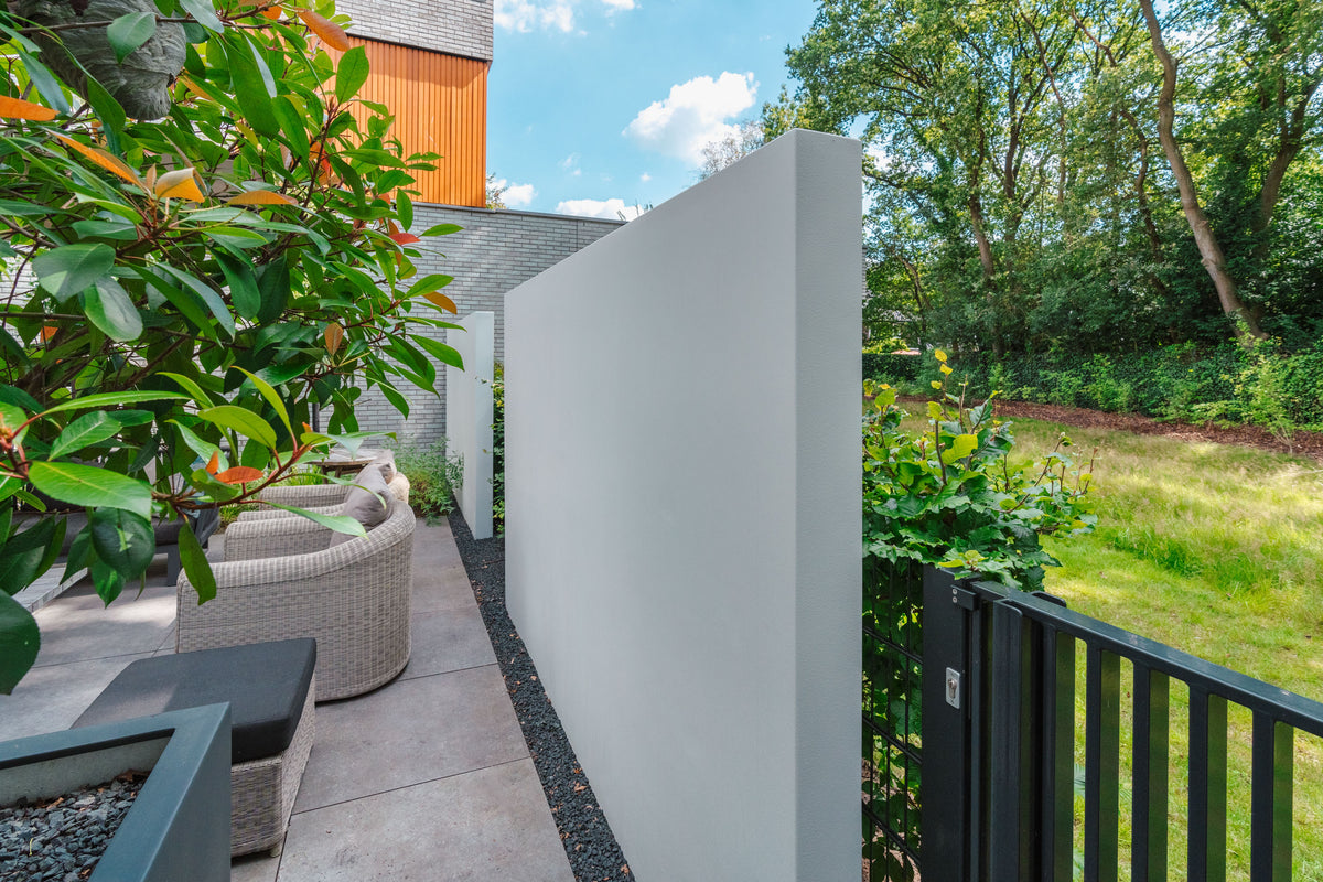 A modern outdoor patio area with a tall white privacy screen, wicker patio seating, a black railing, and a lush green garden background under a blue sky.